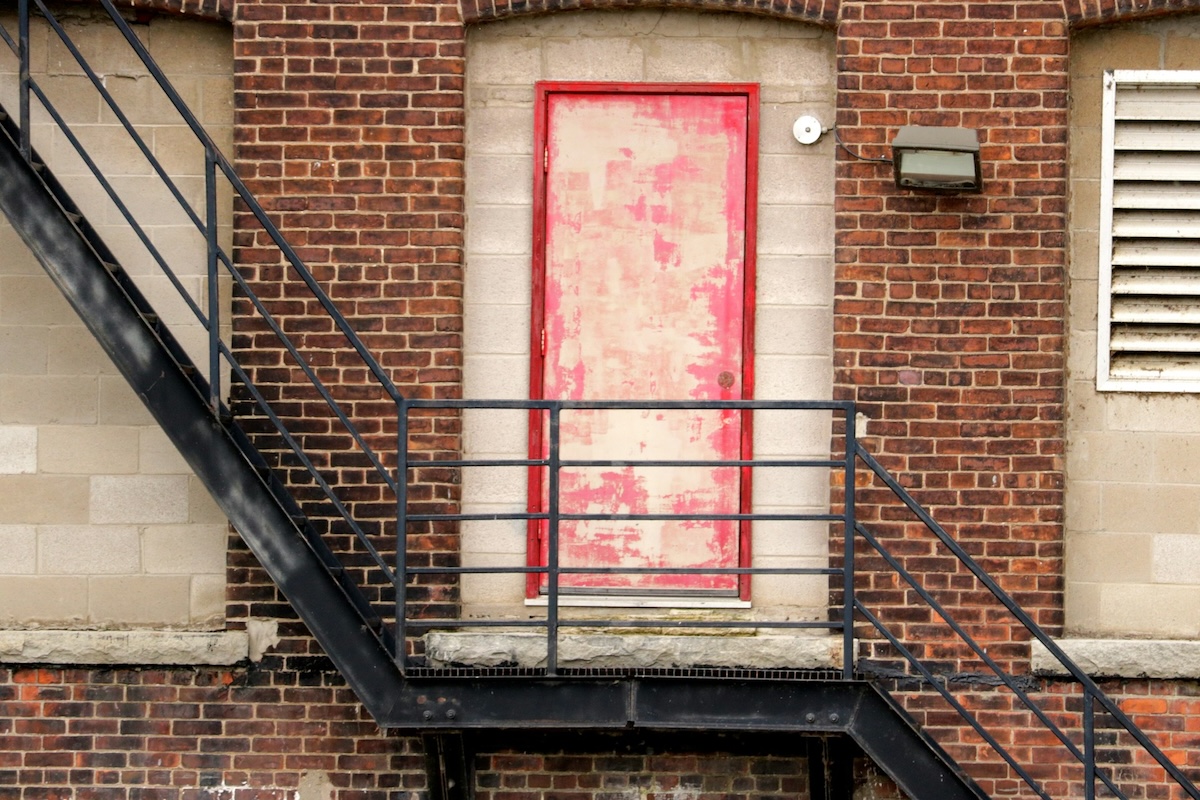 Photo centered on a single, weather-worn apartment door set into a brick wall halfway up a metal staircase